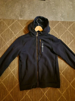 Marinblå hoodiejacka Sail Racing - Mörkblå hoodiejacka från Sail Racing med dragkedja framtill och bröstficka med logga. Jackan har huva med snörning och är tillverkad i mjukt bomullsmaterial. Perfekt för dig som gillar sportig och clean stil. Storleken är 160 och passar för XS/S.