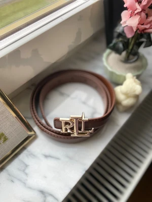 Brunt skärp från Ralph Lauren - Snyggt brunt bälte från Ralph Lauren med RL-logga i guld på spännet. Bältet är gjort i slätt skinn och har stilrena sömmar längs kanterna. Perfekt för att lyfta din outfit med en exklusiv touch och den klassiska RL-detaljen framtill.
