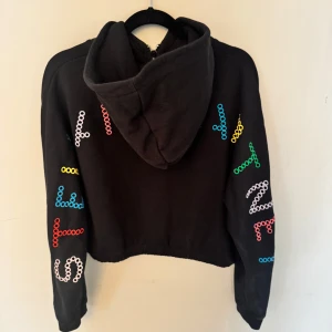 Svart hoodie Stella McCartney Kids - Cool svart hoodie från Stella McCartney Kids med färgglada bokstäver på ärmar och bröst. Croppad modell med huva och dragkedja framtill. Tillverkad i mjuk bomull.