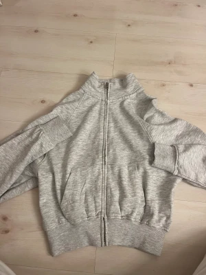 Grå zip hoodie från Zara - Snygg ljusgrå hoodie från Zara i storlek S. Tröjan har hel dragkedja framtill, ribbade muddar och två stora fickor. Perfekt för chill dagar och har en klassisk, enkel look med hög krage. Materialet är mjukt och skönt, troligtvis bomull och polyester.