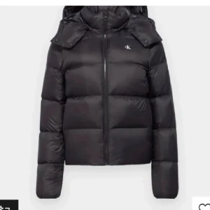 Svart pufferjacka från Calvin Klein - Svart pufferjacka från Calvin Klein med huva och diskret CK-logga på bröstet. Jackan har en quiltad design, dragkedja framtill och långärmade ärmar. Perfekt för kalla dagar när du vill hålla dig varm och samtidigt se snygg ut.