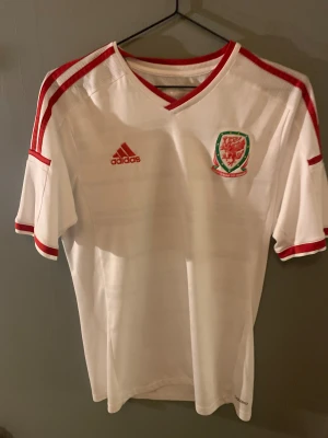 Wales vit fotbollströja Adidas - Säljer en officiell Wales fotbollströja från Adidas i vit polyester med röda detaljer och ränder på axlarna. Tröjan har korta ärmar, V-ringad krage och Wales landslagets emblem på bröstet. Perfekt för dig som vill representera Wales på match eller träning.