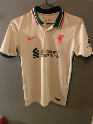 Liverpool FC vit Nike fotbollströja - Liverpool FC matchtröja från Nike i vit polyester med korta ärmar. Tröjan har gröna och orange detaljer på kragen, klubbmärke och Nike-logga i orange samt sponsortryck på bröstet. Klassisk pikékrage med knappar och gröna muddar vid ärmslut.