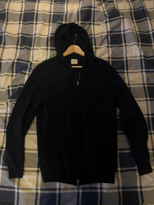 John Henric Kashmir Kofta - Mjuk och lyxig svart hoodie från John Henric i cashmere. Tröjan har huva med snörning, hel dragkedja framtill och långa ärmar. Perfekt för dig som vill ha en stilren och bekväm look med premiumkänsla. Kan gå ner i pris vid snabbköp