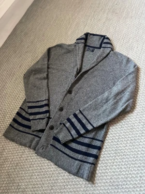 Grå kofta med marinblå ränder - Snygg grå kofta från Watsons med marinblå ränder på ärmar och nederkant. Koftan har knappar framtill, ribbade muddar och en krage med randiga detaljer. Materialet är 60% bomull, 30% polyester och 10% ull, vilket gör den både mjuk och varm.