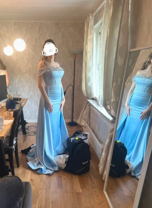 Ljusblå lång balklänning med pärldetaljer - Ljusblå långklänning med offshoulder och glittriga pärldetaljer över bysten. Längst ner finns små fläckar som går att tvätta bort. Pris kan diskuteras! Klänningen har inbyggda sömmar och nålar (inget du kommer känna av) som gör att den kan sys ut några centimeter vid midjan vid behov!