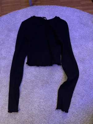 Svart croppad långärmad topp FB Sister - Säljer en svart croppad långärmad topp från FB Sister i storlek XS. Toppen är ribbad med vågig kant vid ärmslut och nederkant, vilket ger en snygg detalj. Perfekt för dig som gillar stilrena och trendiga plagg.