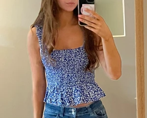 Blommig blå croppad topp från Pigalle - Säljer en söt croppad topp från Pigalle i blått med vitt blommigt mönster. Toppen har breda axelband, smockad midja och volangkant nedtill. Perfekt för varma dagar och ger en fräsch vibe till jeans eller kjol.