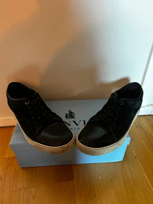Svarta sneakers från Lanvin Paris - Snygga svarta sneakers från Lanvin Paris med platt vit sula och rund tå. Ovandelen är i mocka med detaljer i blankt skinn på tån och snörning framtill. Perfekt för dig som gillar stilrena och lyxiga sneakers med exklusiv känsla. De har en skada på undersidan på ena skon, men inget som påverkar användning och inget som syns. 