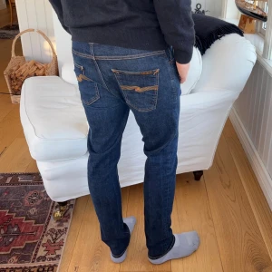 Nästan nya Mörkblå jeans från Nudie Jeans - Säljer ett par mörkblå jeans från Nudie Jeans med klassisk orange söm och snygg broderad detalj på bakfickorna. Modellen har normal passform och raka ben, tillverkade i slitstarkt bomullsmaterial. Perfekt för dig som gillar stilrena och tidlösa jeans.