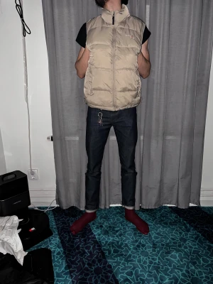 Beige dunväst från Outdoor - Snygg beige dunväst från Outdoor med hög krage och dragkedja framtill. Västen har två stora fickor på sidorna och quiltad design som ger en puffig look. Perfekt att slänga över en hoodie eller t-shirt för extra värme. Neutral färg som passar till allt.