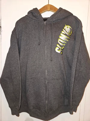 Slednecks hoodie med tryck - Mörkgrå hoodie från Slednecks med fet tryck i vitt och neongult både fram och bak. Bra skick. Strl M. Mått: Längd 70 cm Bredd 57 cm Ärmlängd 56 cm