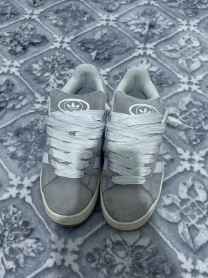 Grå Adidas Campus sneakers strl 43 - Säljer ett par grå Adidas Campus sneakers med klassiska vita ränder och logga på tungan och hälen. Skorna har rund tå, platt sula i gummi och snörning framtill. Ovandelen är i mocka och insidan är mjukt fodrad för extra komfort. Perfekt för dig som gillar stilrena och tidlösa sneakers.