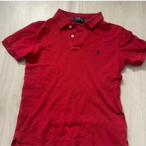 Röd pikétröja från Ralph Lauren - Klassisk röd pikétröja från Ralph Lauren med blå broderad logga på bröstet. Tröjan har korta ärmar, krage och två knappar framtill. Tillverkad i bomull och har en normal passform. Perfekt för en stilren och sportig look. Strl xs Pris kan diskuteras 