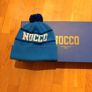 Blå NOCCO mössa med tofs - Snygg blå mössa från NOCCO med vit logga framtill och mörkblå ränder. Mössan har en stor mörkblå tofs på toppen och bred uppvikt kant. Perfekt för dig som gillar sportiga accessoarer och vill sticka ut med en cool look. Pris kan diskuteras. Boxen kommer med.