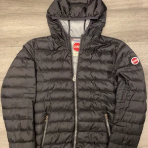 Svart pufferjacka från Colmar - Svart pufferjacka från Colmar med huva och två sidofickor med dragkedja. Jackan har ett quiltat mönster och Colmar-logga på ärmen. Perfekt för kyliga dagar och har en snygg, sportig look. Materialet är syntet och jackan är långärmad. Defekter pytte små hål vid fickorna som knappt syns s men liten i storleken 