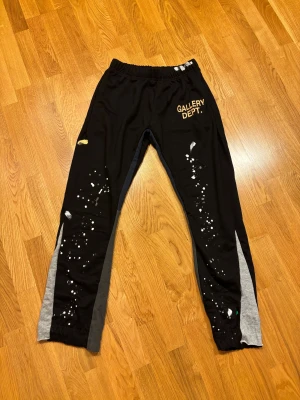 Svarta Gallery Dept mjukisbyxor L - Svarta mjukisbyxor från Gallery Dept i storlek L. Byxorna har elastisk midja, grå och mörkgrå paneler längs benen och vitt stänkmönster framtill. Logotypen 'GALLERY DEPT.' i gult på vänster lår. Tillverkade i 100% bomull för skön känsla.