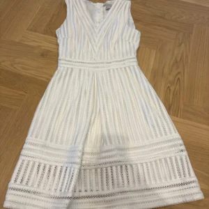 Vit kortklänning med spets från H&M - Supersöt vit kortklänning från H&M med v-ringning och ärmlös design. Klänningen har ett randigt spetsmönster och markerad midja som ger en snygg siluett. Perfekt för sommarens alla tillfällen! Materialet är bomull och polyester.