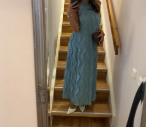 Blå långklänning med fransdetaljer - Säljer en blå långklänning med hög hals och ärmlös design. Klänningen har unika vågiga fransdetaljer över hela tyget som ger en cool och trendig look. Passformen är normal och materialet känns lätt och luftigt, perfekt för sommarens alla tillfällen. För referens är jag 163 cm. Storlek 34/XS. Använd en gång