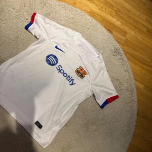 Vit FC Barcelona fotbollströja Nike - Säljer en vit FC Barcelona fotbollströja från Nike med Spotify-logga på bröstet och klubbmärke broderat. Tröjan har korta ärmar med blå och röda detaljer vid ärmslut. Perfekt för dig som vill visa ditt stöd för Barca.