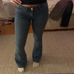 Blå bootcut jeans med hög midja - Snygga blå jeans med bootcut passform och hög midja. Klassisk femficksmodell med knapp och dragkedja framtill. Jeansen har en smickrande siluett som sitter tajt över låren och släpper ut vid vaden. Perfekta för dig som gillar retrovibbar och bekväma jeans.