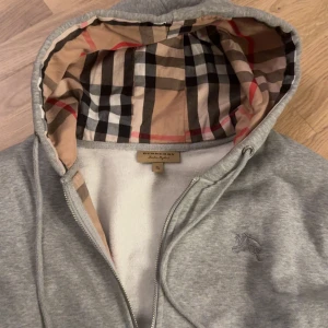 Grå Burberry hoodie med dragkedja - Säljer en grå hoodie från Burberry med klassiskt rutigt foder i huvan och broderad logga på bröstet. Tröjan har dragkedja framtill och snörning i huvan. Perfekt för dig som vill ha en stilren och lyxig vardagslook.