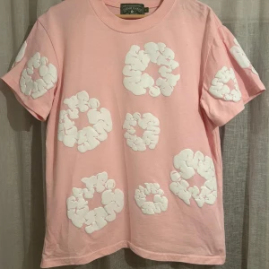 Rosa t-shirt med vita 3D-mönster Denim Tears - Säljer en rosa t-shirt från Denim Tears med coola vita 3D-mönster över hela plagget. T-shirten har rund hals och korta ärmar. Materialet är mjuk bomull och passformen är normal. Perfekt för dig som gillar unika och trendiga plagg.