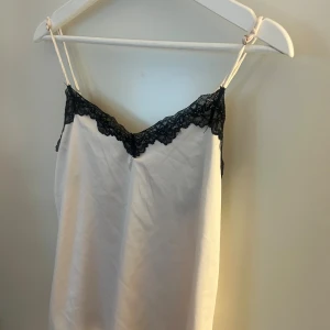 Vitt linne med spets från Primark - Snygg vitt linne från Primark med tunna axelband och svart spetsdetalj längs v-ringad hals. Linnet har en rak och enkel siluett som ger en chill vibe, perfekt för dig som gillar minimalistisk stil med en touch av spets.