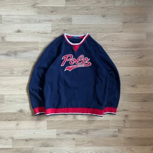 Mörkblå Polo Ralph Lauren tröja - Snygg mörkblå tröja från Polo Ralph Lauren med stort rött och vitt Polo-tryck på bröstet. Tröjan har långa ärmar, ribbade muddar och krage i rött och vitt. Tillverkad i en mjuk blandning av polyester och bomull, perfekt för chill dagar. Klassisk och sportig look.  Bilder på måtten finns tillgängliga. Fråga gärna! Snabb leverans!
