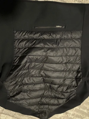 Svart pufferjacka från Parajumpers - Svart pufferjacka från Parajumpers med quiltad framsida och släta, långärmade sidor. Jackan har en gul detalj vid halsen, bröstficka med dragkedja och broderad logga på ärmen. Perfekt för dig som gillar stilren och sportig look.