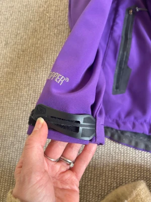 Lila vindjacka från The North Face - windbreaker - Snygg lila vindjacka från The North Face med huva och grått fleecefoder. Jackan har dragkedja framtill, två sidofickor med dragkedja och en bröstficka. Justerbar huva med snörning och broderad logga på bröstet. Perfekt för blåsiga dagar och outdoor.