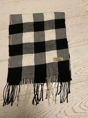Rutig svartvit halsduk från Burberry - Snygg halsduk från Burberry i klassiskt rutigt mönster med svarta, vita och gråa rutor. Gjord av 100% lammull och har fransar i ändarna. Perfekt för att ge din outfit en cool och tidlös vibe. Märkt med Burberry London-tag.