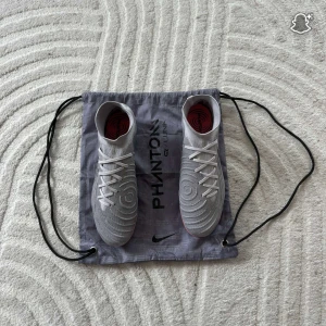 Nike Phantom GX Luna fotbollsskor - Nike Phantom GX Luna fotbollsskor i ljusgrå färg med snygga mönstrade detaljer och svart swoosh. Skorna har snörning och en högre strumpliknande krage för extra stöd. Sulan är iriserande med dobbar för gräsplan. Perfekt för dig som vill sticka ut på planen. Inte använda många gånger kanske tre, fyra gånger 