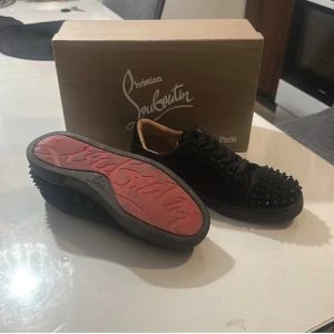 Christian Louboutin svarta sneakers med nitar - Svarta sneakers från Christian Louboutin med klassisk röd sula och coola svarta nitar över tå och häl. Skorna har snörning och är tillverkade i mocka och skinn. Medföljer extra nitar. Perfekt för dig som vill sticka ut med en edgy och lyxig stil.