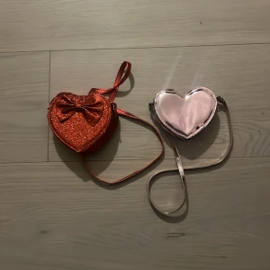 Axelväska, barn - Säljer två hjärtformade barnaxelväskor med lång rem. Ena väskan är röd med glittrig framsida och en stor rosett, den andra är rosafärgad med metallic-ytan. Perfekt för barn som gillar unika accessoarer med lite extra bling och cool form.