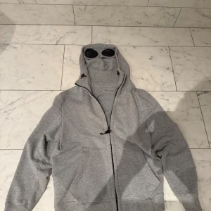 Grå hoodie med goggles från C.P. Company - Säljer en grå hoodie från C.P. Company med ikoniska goggles i huvan. Jackan har dragkedja framtill, snörning vid huvan och två stora fickor. Materialet är mjuk bomull och passformen är normal. Perfekt för dig som vill sticka ut med streetwear-vibe. Pris går att diskutera 