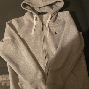 Grå hoodie från Polo Ralph Lauren - Snygg grå hoodie från Polo Ralph Lauren med dragkedja och huva. Tröjan har vita snören, ribbade muddar och den klassiska broderade loggan på bröstet. Perfekt för chill dagar och enkel att matcha med jeans eller joggers.