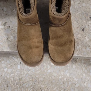 Bruna UGG boots i mocka - Säljer ett par klassiska UGG boots i brun mocka med mjukt, fluffigt foder på insidan. Skorna har rund tå, platt sula och är låga i modellen. Perfekta för kalla dagar och har en enkel, tidlös look med UGG-logga bak.