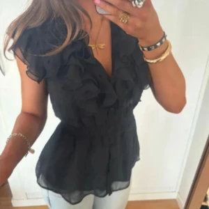 Svart blus med volang och v-ringning - Säljer en svart blus med v-ringning och stora volanger framtill. Blusen är ärmlös och har ett lätt transparent material som ger en luftig känsla. Perfekt för dig som gillar romantiska detaljer och vill ha en snygg topp till jeans eller kjol.