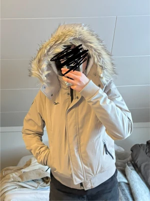 Beige vinterjacka med fuskpäls från Hollister - Säljer en beige vinterjacka från Hollister med fluffig fuskpäls på huvan och mjukt vitt teddyfoder. Jackan har dragkedja, knappar och två fickor med dragkedja. Snygg, varm och perfekt för kalla dagar. Stretchmaterial för extra komfort.