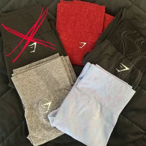 Gymshark leggings i olika färger - Säljer flera par Gymshark leggings i snygga färger: röd med mönster, svart med vågigt mönster och ljuslila. Alla har hög midja och är tillverkade i stretchigt syntetmaterial som sitter skönt och följer kroppen. Priser: Gråa: 300kr,  Mönstrade (svart,blå,röd) : 3 st för 1200kr, 2st för 850, 1 för 450 .Gråa är använda ett fåtal gånger men i fint skick. Resterande byxor är helt nya och aldrig använda. 