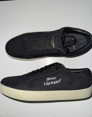 Svarta Saint Laurent sneakers - Svarta sneakers från Saint Laurent med vit kontrastfärgad sula och broderad logga på sidan. Skorna har rund tå, snörning och är tillverkade i canvas med detaljer i skinn. Innersulan är svart med guldfärgad Saint Laurent-text. Snygg och stilren design.