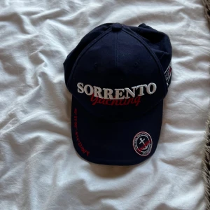 Sorrento Yachting marinblå keps - Marinblå keps från Sorrento Yachting med broderad text i vitt och rött framtill, patch med ankare och detaljer på sidorna. Justerbar baktill med broderad text 'ITALIA' och segelbåt. Perfekt accessoar för en sportig och nautisk look.