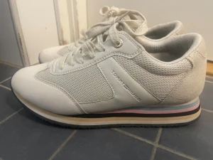 Vita sneakers Tommy Hilfiger pastell - Snygga vita sneakers från Tommy Hilfiger med mesh och mockadetaljer. Skorna har snörning och coola pastellfärgade ränder i rosa, blått och gult på sulan. Klassisk logga bak och diskret branding på sidan. Perfekta för dig som gillar stilrena men ändå unika sneakers.