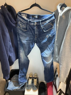 Replay Anbass blå jeans med slitning - Säljer ett par Replay Anbass jeans i blå tvätt med slitningar och hål på ena benet. Klassisk femficksmodell, smal passform och snygga detaljer. Jeansen är gjorda i bomull och har en cool, använd look.