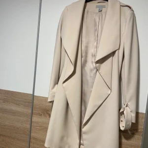 Beige trenchcoat med knytärmar - Stilren beige trenchcoat från H&M med breda slag och knytband vid ärmsluten. Jackan har en mjuk, slät yta och är helfodrad med ett glansigt innerfoder. Perfekt för dig som gillar en klassisk och clean look.