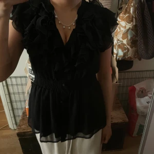 Svart blus med volanger och knappar - Säljer en svart blus med volanger längs hela framsidan och v-ringad hals. Blusen har korta ärmar, knappar framtill och smockad midja som ger en snygg siluett. Materialet är lätt och transparent, perfekt för att skapa en cool och dressad look.