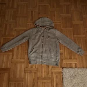 Grå Burberry hoodie med rutig huva/ PRIS KAN DISKUTERAS/ - Snygg grå hoodie från Burberry med klassiskt rutigt mönster på insidan av huvan. Tröjan har dragkedja, snörning vid huvan och broderad Burberry-logga på bröstet. Mjuk insida i fleece och två fickor framtill. Perfekt för chill dagar.