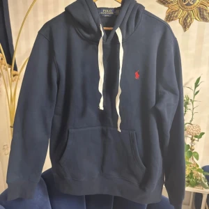 Marinblå hoodie från Polo Ralph Lauren - Snygg marinblå hoodie från Polo Ralph Lauren med vit snörning och röd logga på bröstet. Slim fit-modell med huva och stor magficka framtill. Perfekt för dig som gillar klassisk streetstyle och vill ha en hoodie med stilren look.