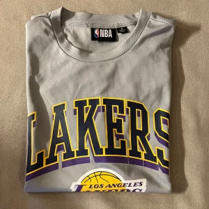 Grå Lakers t-shirt  - Snygg grå Los Angeles Lakers t-shirt med stort tryck i gult, lila och svart på bröstet. Officiell NBA-logga i nacken. Tillverkad i mesh-material som andas, perfekt för sport eller häng. Klassisk rund hals och korta ärmar. Lite stor i storleken som det ska va. Nästan inte använd. 